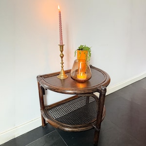 Puede incluir: Una mesa auxiliar de dos niveles, de color marrón oscuro, con un estante inferior tejido. Una vela rosa retorcida encendida está en un candelabro de latón. Un portavelas de cristal con una vela encendida y una pequeña maceta naranja con vegetación están en el estante superior.