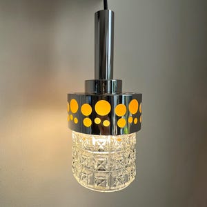 Op de afbeelding: Een chromen hanglamp met een heldere glazen kap en een gele gepatenteerde metalen kap. De lamp is aan en verlicht de kamer.