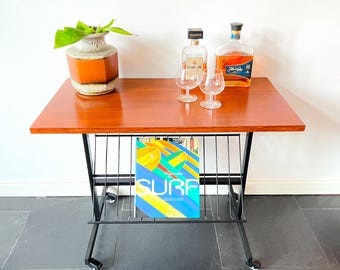 May include: A mid-century modern magazine rack with a wooden top and black metal frame. The rack has a magazine holder and wheels. There are two bottles of liquor and two glasses on the top of the rack. A potted plant is on the left side of the table.