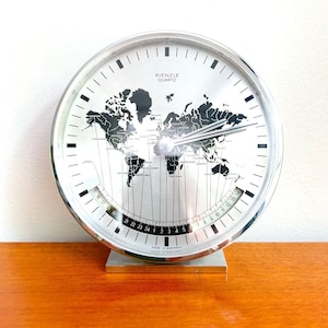 Può includere: Un orologio mondiale argentato con un design di mappa del mondo in bianco e nero. Il quadrante dell'orologio include anche una scala di tempo di 24 ore. L'orologio è realizzato in Germania e riporta la scritta "Kienzle Quartz" sul quadrante.