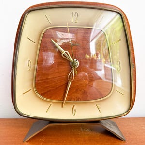 Peut inclure: Une horloge vintage carrée avec un cadran crème et un centre en grain de bois. L'horloge a des aiguilles et des marqueurs dorés, et un cadre en bois marron. Elle repose sur un support en métal. Le cadran est protégé par un verre transparent.