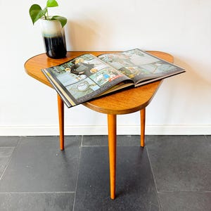 Op de afbeelding: Een driehoekige houten bijzettafel met drie taps toelopende poten en een gladde houtnerf. Een open stripboek met kleurrijke illustraties ligt plat op het oppervlak, vergezeld door een kleine keramische vaas met een wit en donkerblauw kleurverloop.