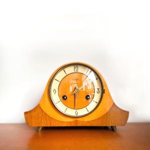 Op de afbeelding: Een vintage houten mantelklok met een lichtoranje wijzerplaat en een heldere glazen afdekking. De klok heeft een gebogen, boogvormig houten frame en goudkleurige metalen poten. De wijzerplaat heeft zwarte cijfers en wijzers.