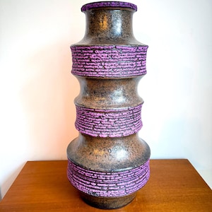 May include: A tall, brown ceramic vase with a textured surface and a cylindrical shape. The vase features bands of bright purple around the middle and top, creating a striking contrast. The vase is a decorative item.