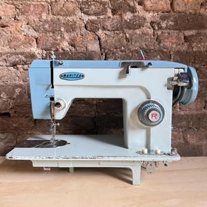 May include: A light blue and off-white Maritza sewing machine featuring a prominent circular dial with a red R symbol, a metal needle assembly, and a flat base plate. The machine has a horizontal top section and a vertical body with mechanical controls.