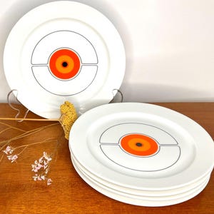 May include: Set of six white ceramic dinner plates featuring a geometric design in orange, black, and white. The design incorporates a circle with a smaller circle inside, surrounded by two concentric rings.