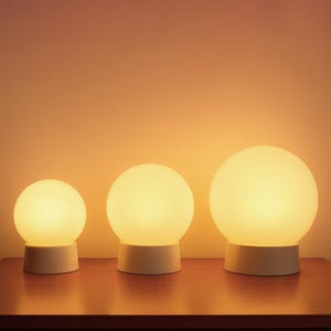 Op de afbeelding: Drie bolvormige tafellampen die een warm licht uitstralen. Elke lamp heeft een ronde, ondoorzichtige kap en een cilindrische voet. De lampen staan op een houten oppervlak tegen een oranje gradiënt achtergrond.