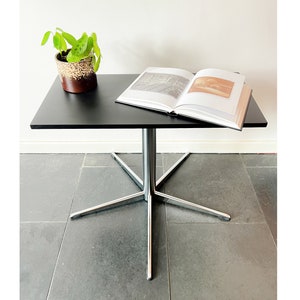Op de afbeelding: Een zwarte rechthoekige tafel met een centrale zilverkleurige metalen voet met vier uitlopende poten. Op het tafelblad ligt een open boek met witte pagina's en een kleine kamerplant in een bruin met crème gespikkelde keramieken pot.