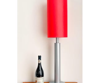 May include: A tall table lamp featuring a cylindrical red fabric shade atop a silver metal post and a circular base. Next to the lamp stands a dark glass wine bottle with a white label that reads Fontanafredda Barolo in black text.