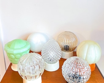 May include: A collection of eight vintage glass lampshades in various shapes and colors, including clear, frosted, and green. The lampshades are all round or oval and have different textures, such as ribbed, textured, and smooth.
