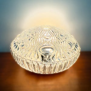 May include: A clear, decorative glass bowl with an intricate, swirling pattern on the top and a ribbed base. The bowl is illuminated from within, creating a soft, warm glow. The bowl is approximately 8 inches in diameter.