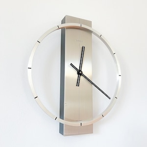 Può includere: Un orologio da parete argentato con un quadrante circolare e lancette nere. L'orologio ha una cornice rettangolare con la scritta "JUNGHANS" stampata su di essa.