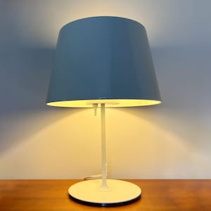 May include: A white table lamp with a light blue lampshade. The lamp is switched on and casting a warm glow.