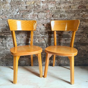 May include: Two matching chairs constructed from light-toned wood with a smooth, polished finish. Each chair features a curved backrest, a rounded seat, and four straight, sturdy legs. The wood grain is visible across the surfaces of the chairs.