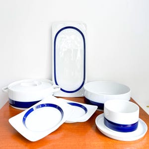 May include: A collection of white porcelain tableware with a bold blue stripe design. The set includes a serving platter, a covered casserole dish, bowls, plates, and a cup with a saucer. The clean, modern aesthetic is suitable for everyday use or special occasions.