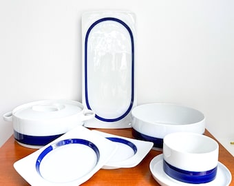 Może przedstawiać: Zestaw białej porcelanowej zastawy stołowej z odważnym, granatowym wzorem w paski. Zestaw zawiera półmisek do serwowania, garnek z pokrywką, miski, talerze i filiżankę ze spodkiem. Czysta, nowoczesna estetyka nadaje się do codziennego użytku lub na specjalne okazje.