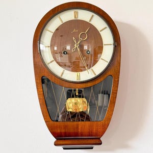 Könnte beinhalten: Eine Vintage-Wanduhr mit Holzrahmen und cremefarbenem Zifferblatt. Die Uhr hat goldfarbene Zeiger und Stundenmarkierungen, mit dem Wort "Jkai" auf dem Zifferblatt. Der untere Teil hat eine Glasscheibe mit einem Pendel.