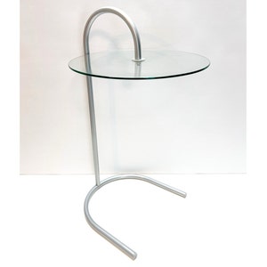 May include: A silver metal side table with a round glass top. The table has a curved design and a single leg.
