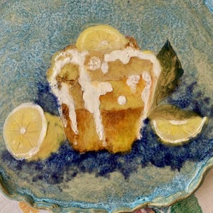May include: A ceramic plate with a light blue, textured surface and a scalloped edge. The plate features a painted lemon cake with white icing, lemon slices, and green leaves. The plate is approximately 12 inches in diameter.