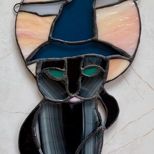 May include: A stained glass suncatcher featuring a black cat wearing a dark blue witch hat. The cat has green eyes and a pink nose. Behind the cat is a circular background made of peach and white glass panels. A silver chain is attached to the top.