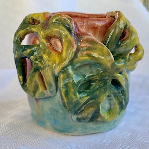 May include: A hand-painted ceramic planter with a unique design. The planter features a cylindrical shape with a light blue base, transitioning to green and yellow leaf-like accents. The interior is a reddish-orange color.