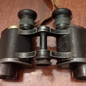 May include: A pair of vintage black binoculars with gold and silver detailing. The binoculars have a leather strap and a textured finish. The lenses are black and the focus knob is textured. The binoculars are on a wooden surface.