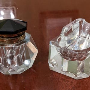May include: Two vintage crystal desk accessories. One is a faceted inkwell with a black lid and gold-toned metal rim. The other is a clear crystal ashtray with a curved design. Both items are on a wooden surface.