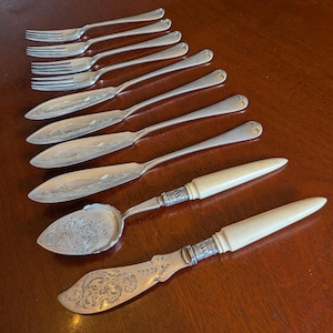 May include: A collection of silver-toned flatware including four forks with simple handles, three fish knives featuring engraved fish motifs, and two serving utensils with ornate scrollwork patterns and smooth off-white handles.