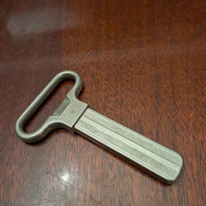 Pode incluir: Um abre-garrafas de metal prateado com um cabo retangular apresentando finas ranhuras verticais paralelas e uma cabeça arredondada de laço aberto, concebida para remover tampas. A ferramenta possui um acabamento mate, liso e robusto.