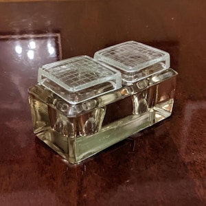 Puede incluir: Un salero doble de vidrio transparente con dos tapas cuadradas con diseño de rejilla. La base rectangular tiene un borde plateado. El salero se encuentra sobre una superficie brillante de color marrón oscuro. Esta pieza vintage es ideal para una cocina o comedor.