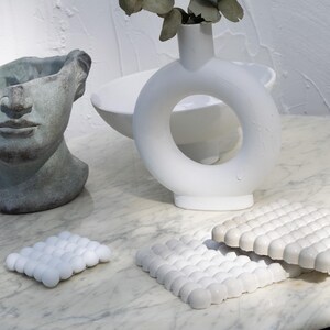Op de afbeelding: Twee witte keramische onderzetters met een textuur, een witte keramische vaas met een ronde opening, een grijze keramische plantenpot in de vorm van een mensenhoofd en een witte keramische kom op een wit marmeren oppervlak.