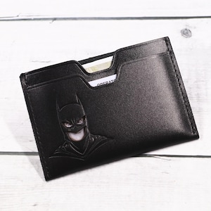 Könnte beinhalten: Schwarzer Lederkartenhalter mit geprägtem Batman-Design. Das Portemonnaie hat mehrere Kartenfächer und ein minimalistisches Design. Ein Teil einer Karte ist in einem der Fächer sichtbar. Der Kartenhalter befindet sich auf einer hellen Holzoberfläche.