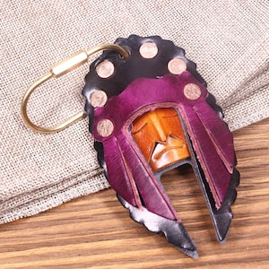 May include: A handcrafted leather keychain featuring a Native American headdress design. The keychain showcases a color palette of black, purple, and tan, with a brass-colored clasp. The leather is intricately cut and shaped, creating a unique accessory.