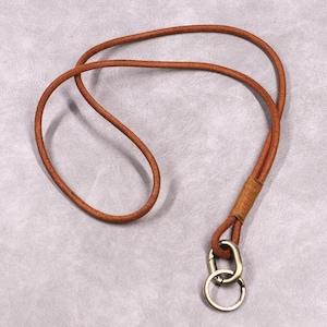 Puede incluir: Un cordón de cuero marrón con un cierre de metal y un llavero. El cordón está hecho de un cordón de cuero redondo y trenzado. Los herrajes metálicos tienen un acabado de latón. El cordón está diseñado para guardar llaves o una tarjeta de identificación.