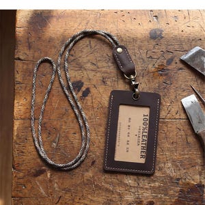 May include: A brown leather ID badge holder with a patterned lanyard. The holder has a clear window and the text "100% LEATHER". The lanyard is a mix of black, white, and grey. The holder is on a wooden surface.