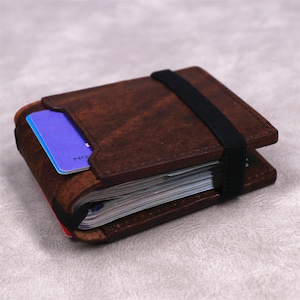 May include: A dark brown leather wallet with a textured finish, featuring a black elastic band wrapped around the center to secure a stack of paper currency and a single blue credit card tucked into the front exterior slot of the wallet.