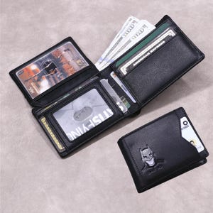 May include: A black bi-fold wallet featuring an embossed Batman logo on the front exterior. The interior includes a clear ID window, multiple card slots, and a compartment containing paper currency. The wallet is constructed from black leather.