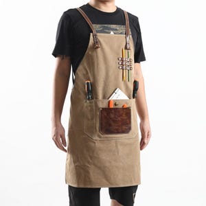 May include: A tan canvas apron with brown leather accents and pockets. The apron has a large front pocket with a leather patch and smaller pockets for tools and pencils. The apron is worn by a person.
