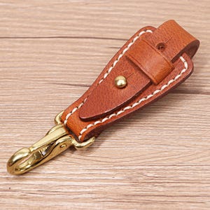 May include: A brown leather key fob with a brass-colored clasp and rivet. The key fob has white stitching around the edges and a small strap with a buckle. The background is a light brown wooden surface.