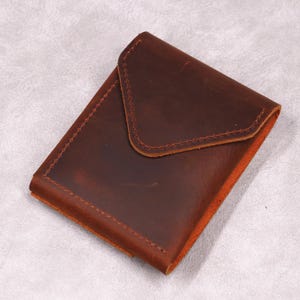 May include: A rectangular, brown leather wallet with a flap closure. The wallet is made of distressed leather and features visible stitching around the edges. The interior is a lighter shade of brown.