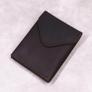 Pode incluir: Uma carteira de couro marrom escuro com fecho de aba. A carteira retangular tem cantos arredondados e uma borda costurada. A aba é presa com uma parte superior curva. O couro tem uma aparência ligeiramente desgastada.