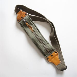 May include: Olive green canvas crossbody bag with a long adjustable strap. Features a full-length zipper, brown leather accents, and metal rivets. The bag is designed with a rectangular shape and a vintage aesthetic.
