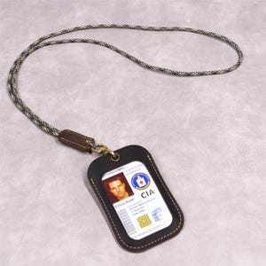 May include: A dark brown leather ID holder with a lanyard. The ID card shows a photo and the text "CIA". The lanyard is a rope-like design with a mix of colors. The holder is attached to the lanyard with a gold-colored clasp.