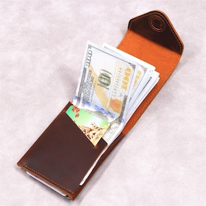 May include: A brown leather wallet, open to reveal a stack of one hundred dollar bills and a credit card. The interior is a lighter shade of brown. The wallet has a simple, classic design.