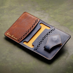 Puede incluir: Una cartera de cuero plegable con un panel exterior marrón y un panel interior azul oscuro con forro amarillo. La cartera incluye bordes perforados decorativos, una pequeña lengüeta de cuero azul y el texto dorado "hillms handmade".