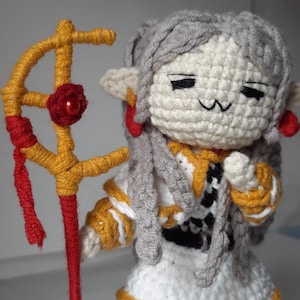 Puede incluir: Una muñeca de crochet con cabello gris, vestida con un vestido blanco con detalles amarillos y negros. La muñeca sostiene un bastón de crochet rojo y dorado.