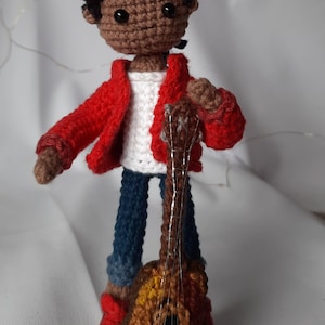 May include: Crocheted doll with brown skin, black hair, and a red jacket holding a brown guitar.