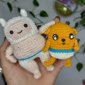 Puede incluir: Dos figuras de ganchillo hechas a mano: una en blanco y rosa claro, la otra en amarillo. La figura blanca tiene una capucha y una cara sonriente. La figura amarilla tiene ojos grandes y un pañal azul y blanco. Ambas se sostienen en una mano.