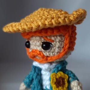 Op de afbeelding: Een gehaakte pop met een gele hoed, oranje baard en blauw jasje. De pop draagt een zonnebloem op zijn borst.
