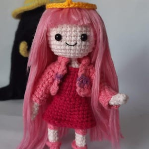 May include: A pink crocheted doll with long pink hair, wearing a red dress, a pink cardigan, and pink boots. The doll has a yellow crown with a blue gem on top.
